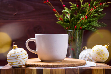 A white cup with a hot drink stands on a wooden board among white Christmas toys next to a green bouquet of fir branches. Christmas Eve concept