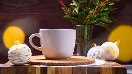 A white cup with a hot drink stands on a wooden board among white Christmas toys next to a green bouquet of fir branches. Christmas Eve concept
