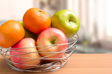 Close-up image of apples and oranges in a wire basket put on tabletop, kitchen blurred in background.