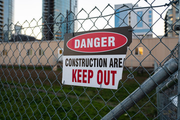 "Danger, keep out, construction area" sign on the chain link fence. Blurred high rise buildings in the background.