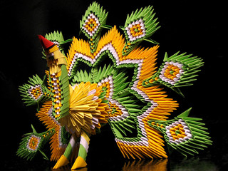 Paper peacock green, yellow and white with a red beak, made in technique of modular origami on a black background. Handmade bird.