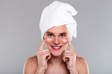 Soft touch. Portrait of handsome shirtless young man in towel on his head applying cream on his face and looking at camera with smile while standing against grey background