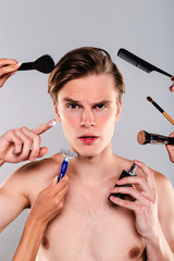 Are you ready for all that in the morning? Handsome young man surrounded by hands holding grooming and cleaning tools looking at camera while standing against grey background