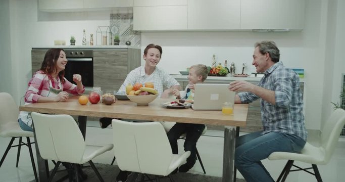 Mature parents with their children spending time on the dinner table watching something on the laptop dad shoo some funny stuff , they have a happy smiling faces in a modern kitchen design.