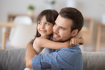 Head shot diverse preschool daughter sitting on sofa together with caucasian young father and hugging. Support protection and warm relations between dad and kid, daddy is best friend concept