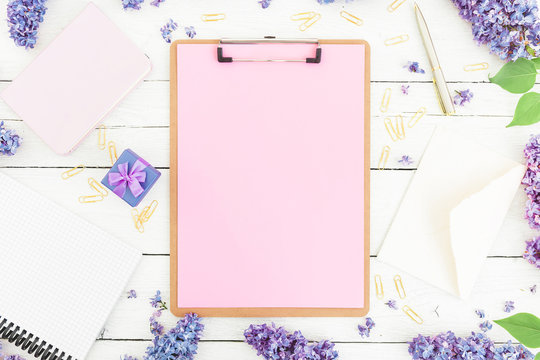 Feminine work place composition with clipboard, notebook, lilac flowers, gift box and accessories on rustic background. Flat lay, top view.