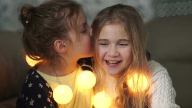Two little girlfriends share secrets. Girl whispering in her sister's ear, both laughing merrily, christmas evening, new year's garland
