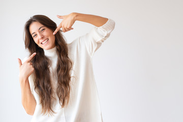 Happy Latin girl pointing at toothy smile and looking at camera. Cheerful young woman with long hair in white sweater. Isolated on white with empty space. Positivity concept