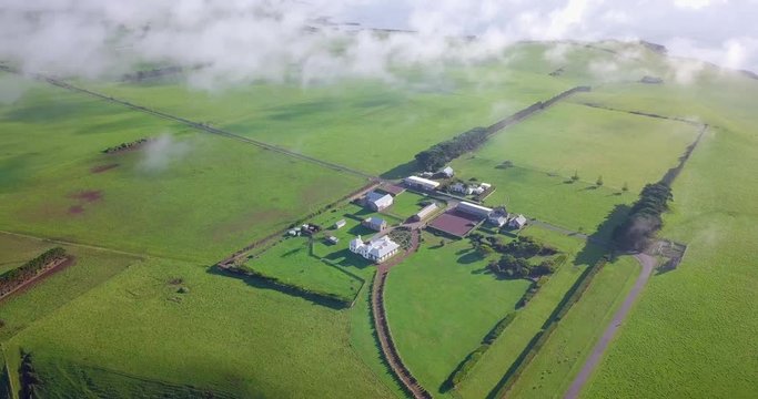 Aerial flight over green field and farmhouse above clouds in Tasmania, Australia