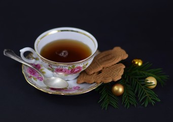 Tasse Tee in einer eleganten Porzellantasse mit Silberlöffel, Spekulatius-Plätzchen, Tannenzweigen und goldenen Christbaumkugeln vor dunklem Hintergrund