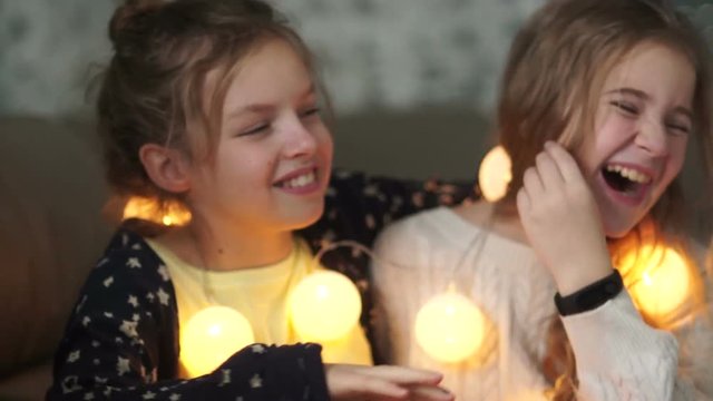 Schoolgirls have a pajama party at home and share secrets. Girl whispering in her sister's ear, both laughing merrily, christmas evening, new year's garland