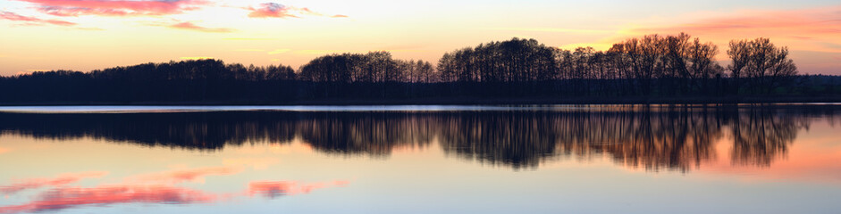 Panorama of a pink and orange sunset soft clouded sky, reflected in the calm water of a rural lake with a silhouetted trees coastline.