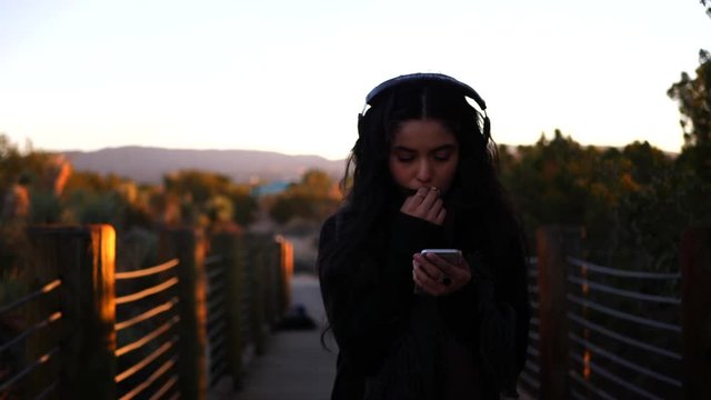 A cute sad girl in slow motion while reading an upsetting text message on her smartphone and listening to music on her headphones at sunset.