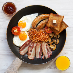 Full English breakfast in a pan with fried eggs, bacon, sausages, beans, toasts and orange juice on white wooden surface, overhead view. Flat lay. From above, top view.