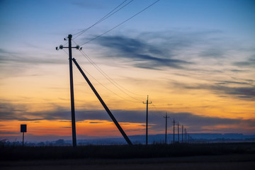 Fototapeta na wymiar Power lines in field on sunrise background. Silhouettes of poles with wires at dawn. Cables of high voltage on warm orange blue sky. Power industry at sunset. Multicolored picturesque vivid sky.