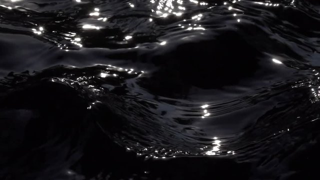 Slow motion of standing waves on black water surface in sun shine reflection. Amazing abstract natural contrast background. High speed camera shooting. Bright play of light on calm ripple.
