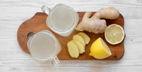 Homemade ginger tea with lemon in glass jars on wooden board over white wooden surface, overhead view. Flat lay, from above, top view.