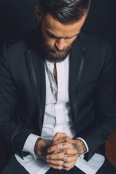 Young man wearing suit, sitting with hands clasped, tattoos on hands, pensive expression