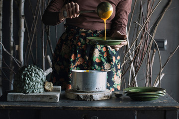 Young woman ladling fresh soup from saucepan at rustic kitchen counter, mid section