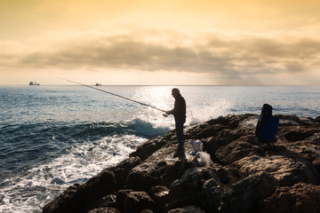 A fisherman standing with the fishing rod in his hand, he is on the rocks at sunset, near him buckets and equipment in bags, on the bottom an oil tanker sails on the horizon, the sky is cloudy and gol © Maurizio Milanesio