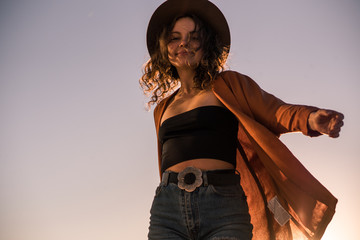 Curly chestnut-haired sexy girl in jeans shorts and hat is spinning and have fun at sunset