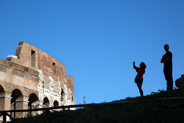 A man and a woman stand on a hill and photograph the Colosseum.