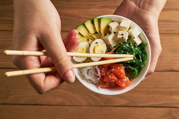 female hands holding chopsticks poke bowl with salmon, avocado rice, Chuka Salad, cheese, quail eggs sprinkled with white and black sesame soy sauce isolated on wooden background top view