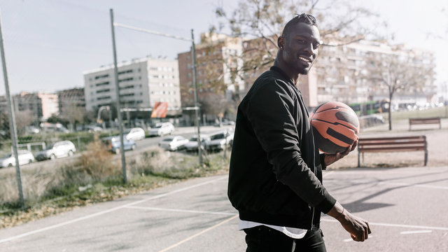 Side view of young african man smiling and holding a ball in a basket on a basketball court