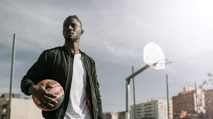 View from below of young african man with black jacket holding a basket ball in a basketball court