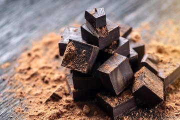 Pieces of dark bitter chocolate with cocoa powder on dark wooden background. Concept of confectionery ingredients