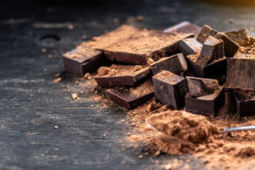 Pieces of dark bitter chocolate with cocoa powder on dark wooden background. Concept of confectionery ingredients