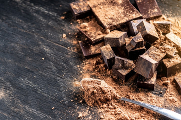 Pieces of dark bitter chocolate with cocoa powder on dark wooden background. Concept of confectionery ingredients