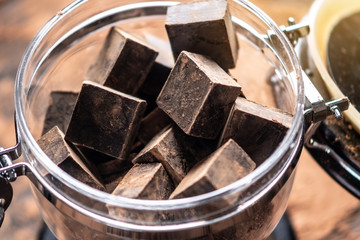 Pieces of dark bitter chocolate with cocoa in a glass jar on wooden background. Concept of confectionery ingredients