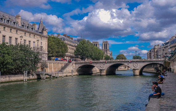 Seine river docks, Paris, France - July 30, 2018: View to the bottom of the Notre Dame towers and bridge crossing the river Seine from a dock with people sitting on the edge of the pier