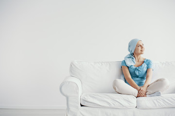 Pretty young woman suffering from breast cancer, wearing blue headscarf and siting cross leg on the couch at home before treatment, copy space on empty wall