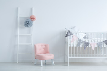 Elegant kid's room with white wooden ladder with grey and pastel pink pompons, small cute armchair and crib with star shape pillows, copy space on empty white wall