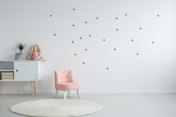 Pastel pink armchair next to wooden cabinet with books, toy and green plant in grey material pot, copy space and golder stars stickers on empty white wall, round carpet on the floor