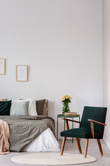 Armchair next to table with flowers in white bedroom interior with posters above bed. Real photo