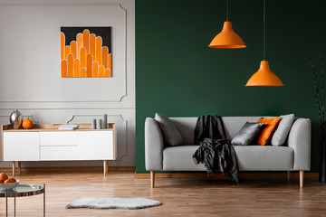 Orange lamps above grey couch in black living room interior with poster above cabinet. Real photo
