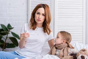Naklejka premium mother taking care of sick daughter in bedroom and checking her temperature with thermometer, looking at camera