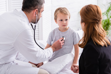 pediatrist in white coat examining sick boy with stethoscope at home
