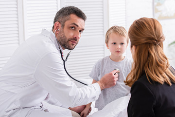 Fototapeta premium pediatrist in white coat examining sick boy with stethoscope and looking at camera at home