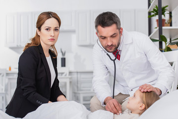 pediatrist in white coat examining sick kid with stethoscope at home, mother looking at camera