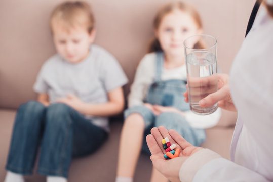 cropped image of pediatrist holding pills and glass of water for sick kids in living room