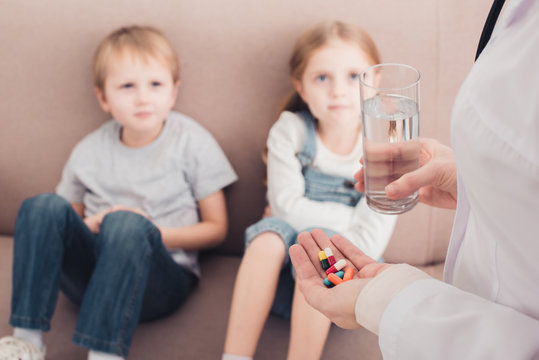 cropped image of pediatrist holding pills and glass of water for sick children in living room