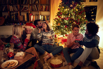 Group of friends sitting next to a Christmas tree, eating Christmas cookies, drinking cocoa and...