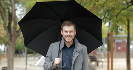 Front view portrait of a happy man walking in a park under the rain holding an umbrella looking at you in winter 