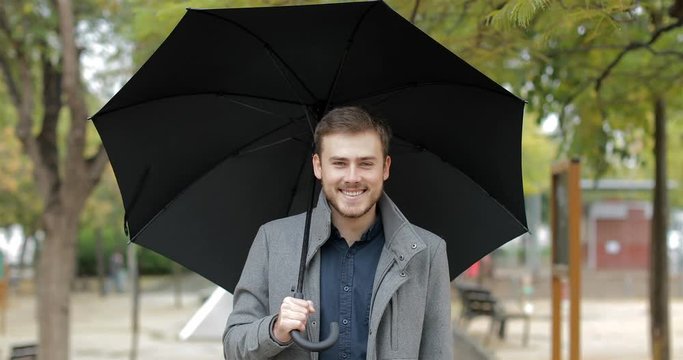 Front view portrait of a happy man walking in a park under the rain holding an umbrella looking at you in winter 