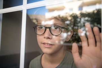 blond child with glasses, he looks calm and serene through the window with his hand resting on the glass. Lights of the sunset are reflected on the glass. Wait for friends to come play with him