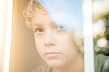 blond child looks pensive and engrossed through the window glass, on the glass reflects the beautiful sunny day and blue sky